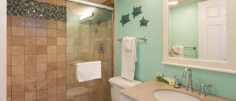 Un baño con ducha de vidrio, inodoro blanco, lavamanos con espejo y decoración de tortugas en la pared.