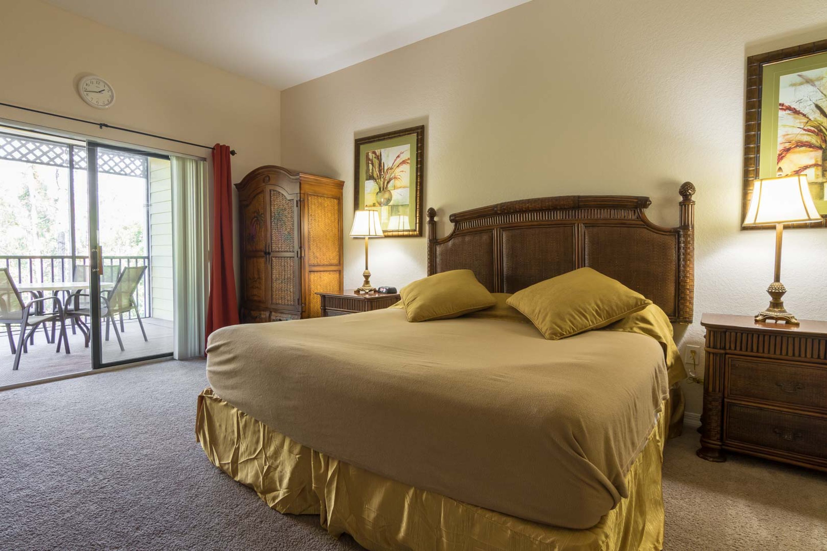 A hotel room with a large bed, a wooden headboard, a nightstand with a lamp, a wardrobe, and a sliding glass door leading to an outdoor area with chairs.