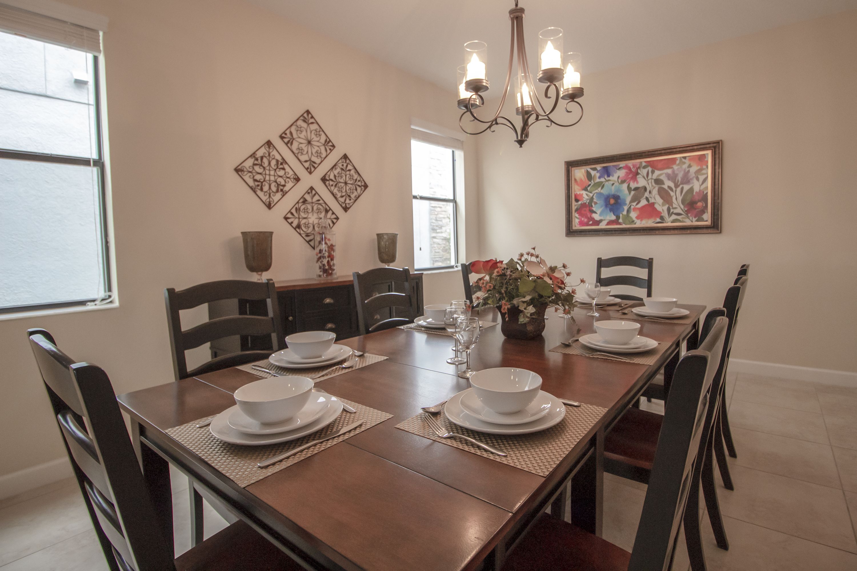 A dining room with a wooden table set for four, surrounded by chairs. There is a chandelier above the table, and artwork is displayed on the walls.