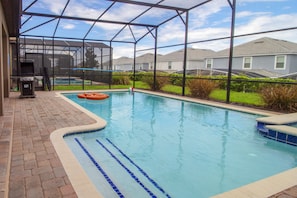 A covered outdoor swimming pool with a grill and lounge chair, surrounded by a fence and houses in the background. - Champions Gate 9027 A (Davenport)