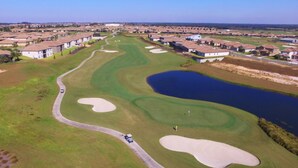 Luftaufnahme eines Golfplatzes mit Sandbunkern, einem Teich und Wohnhäusern im Hintergrund.