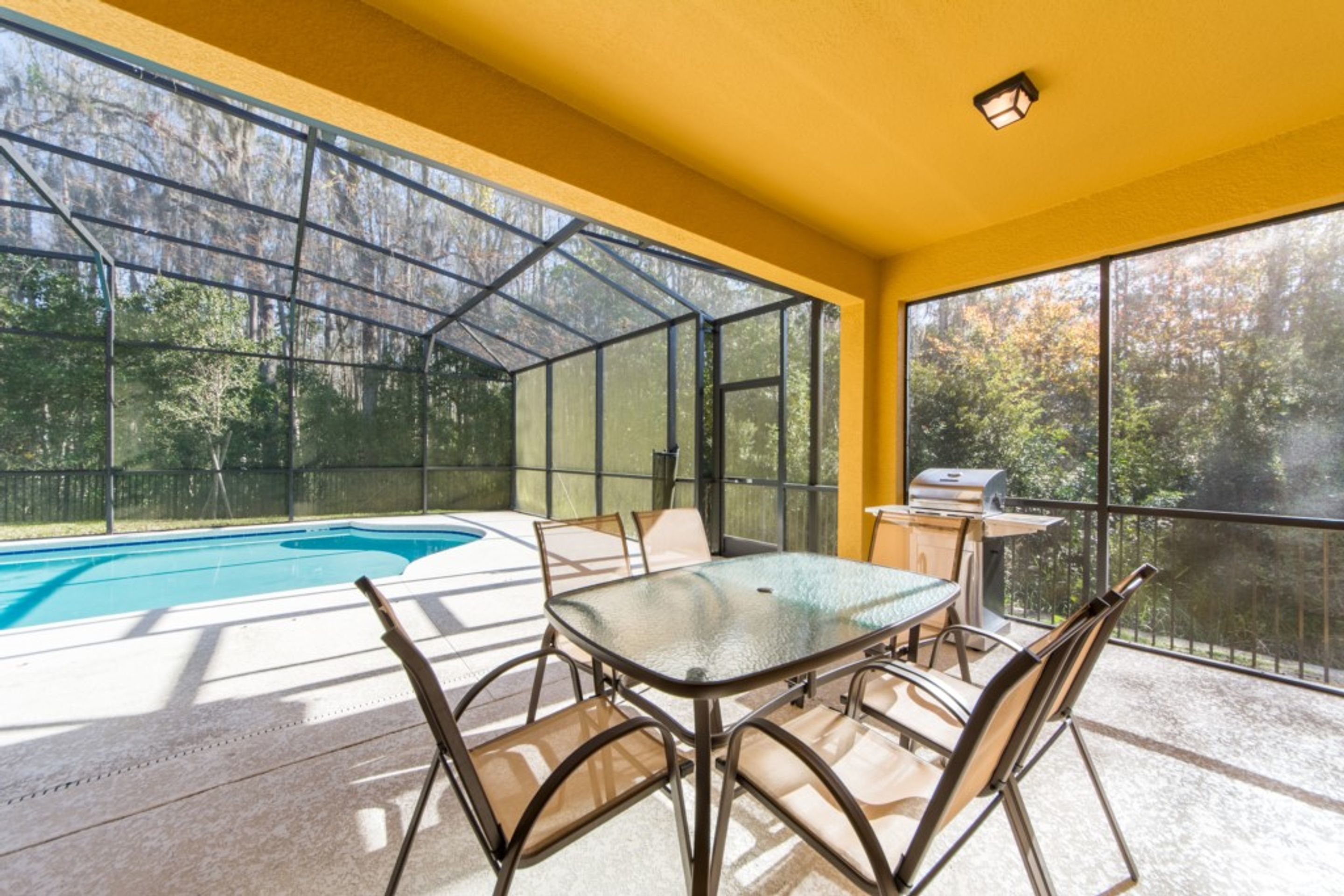 A sunlit patio with a glass table and chairs, a barbecue grill, and a swimming pool.