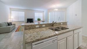 A modern kitchen with granite countertops, a double sink, and white cabinetry. There is a dining area with a sofa and a TV in the background. - Bella Vida 871 (Kissimmee)
