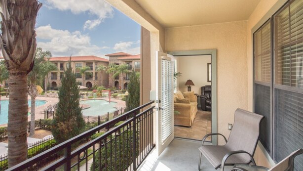 A balcony with a view of a pool and a building with a red roof. There are two chairs and a table on the balcony. - Bella Piazza 904 - 526 (Davenport)