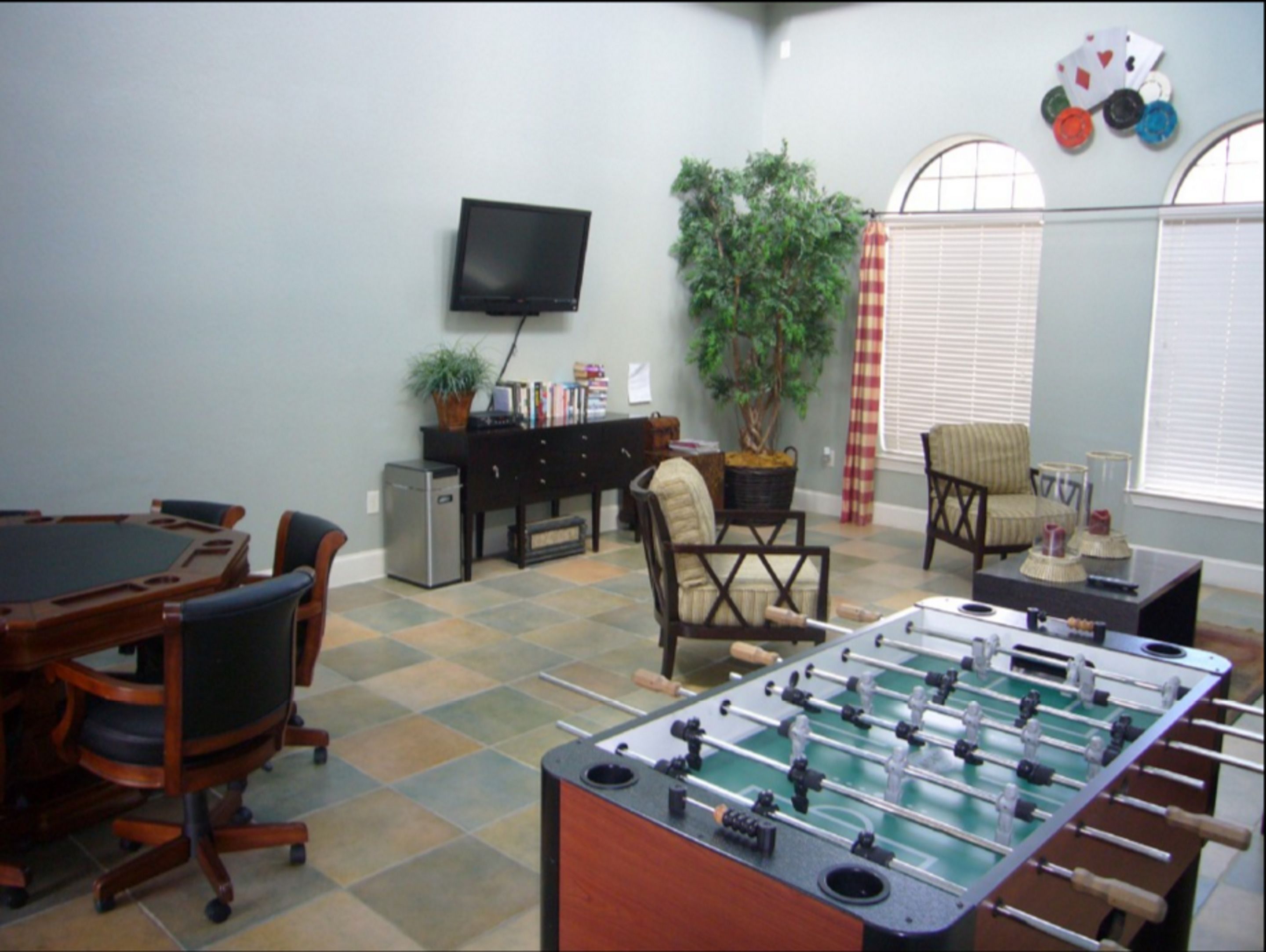A room with a foosball table, a television, a bookshelf, a plant, and a window with blinds.
