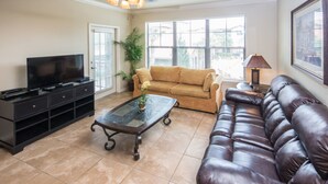 A living room with a television, a glass coffee table, a brown leather sofa, a beige couch, and a potted plant. - Bella Piazza 904 - 526 (Davenport)