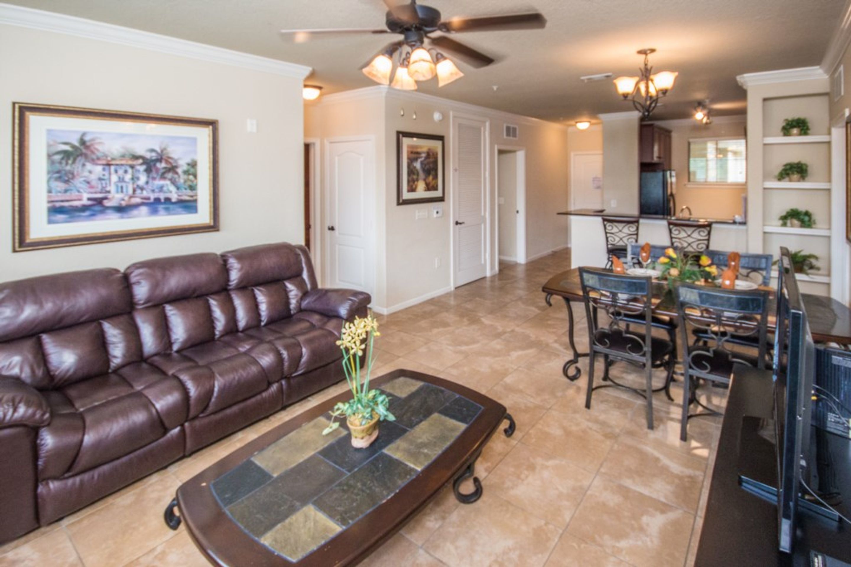 A living room with a brown leather sofa, a glass coffee table with a plant, a ceiling fan, and a dining area with metal chairs and a chandelier.