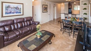 A living room with a brown leather sofa, a glass coffee table with a plant, a ceiling fan, and a dining area with metal chairs and a chandelier. - Bella Piazza 904 - 526 (Davenport)