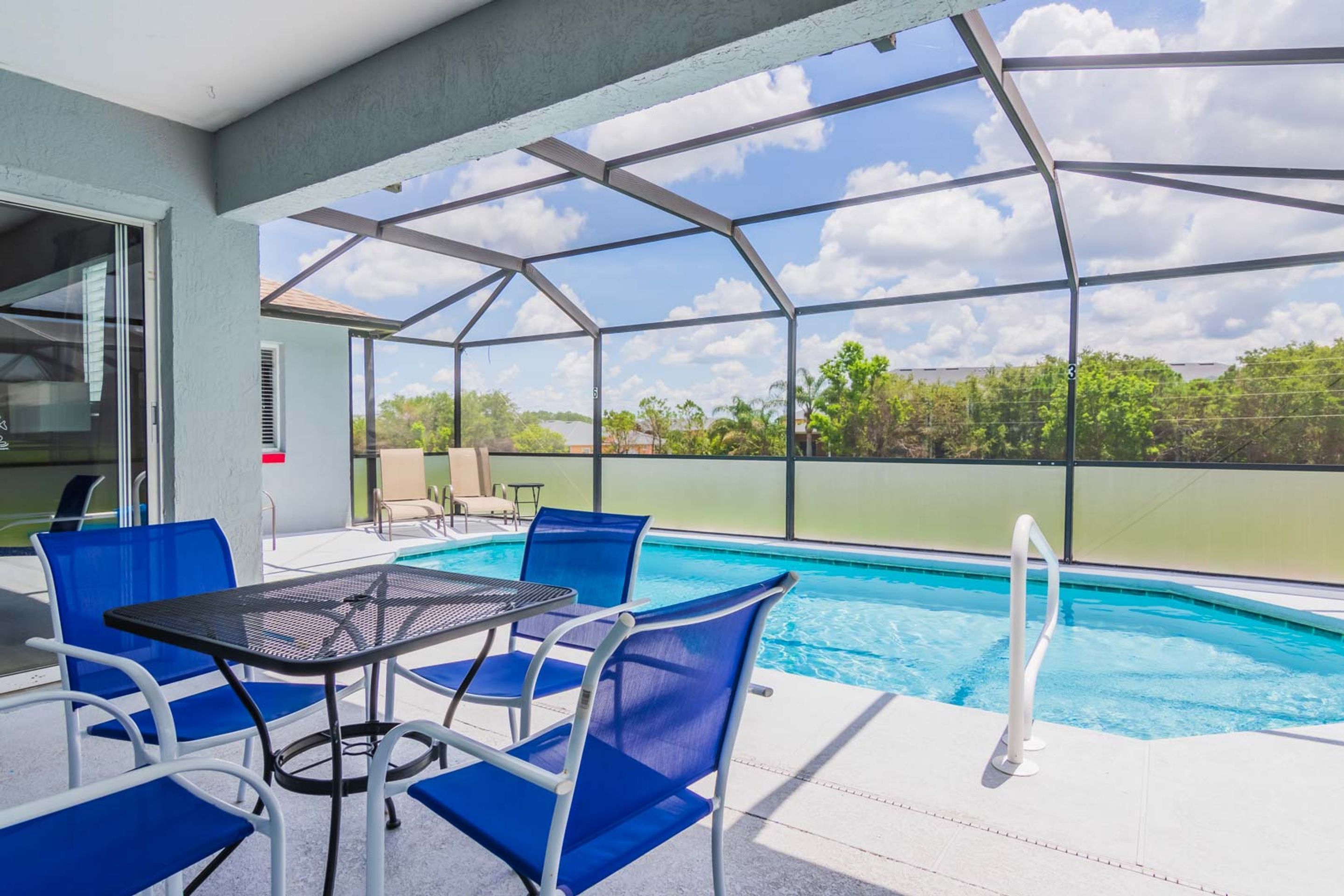 A pool area with blue chairs and a table, a glass enclosure, and a view of greenery.