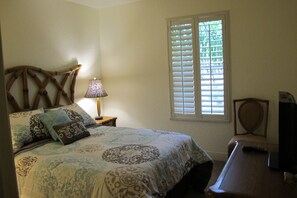 A bedroom with a bed, a nightstand, a lamp, a window with blinds, and a television.