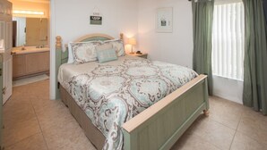 Une chambre à coucher avec un lit, une table de chevet, un ventilateur de plafond, une fenêtre avec des rideaux et une salle de bain visible à travers une porte ouverte.