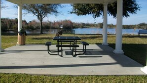 Un espace de pique-nique abrité, équipé d’une table et de bancs, d’une poubelle, et offrant une vue sur un lac bordé d’arbres, avec un banc visible au loin.