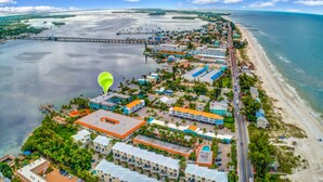 An aerial view of a coastal resort with multiple buildings, a pier extending into the water, and a beach adjacent to the property. - Anna Maria Island 1301 2B (Bradenton Beach)