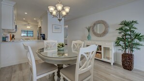 A dining area with a glass table, white chairs, a chandelier, a white cabinet, a mirror, and a potted plant. - Anna Maria Island 1301 2B (Bradenton Beach)