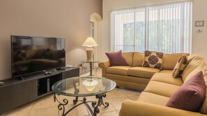A living room with a flat-screen TV, a glass coffee table, a sofa with patterned cushions, and a window with blinds. - Windsor Palms 2200 (Kissimmee)