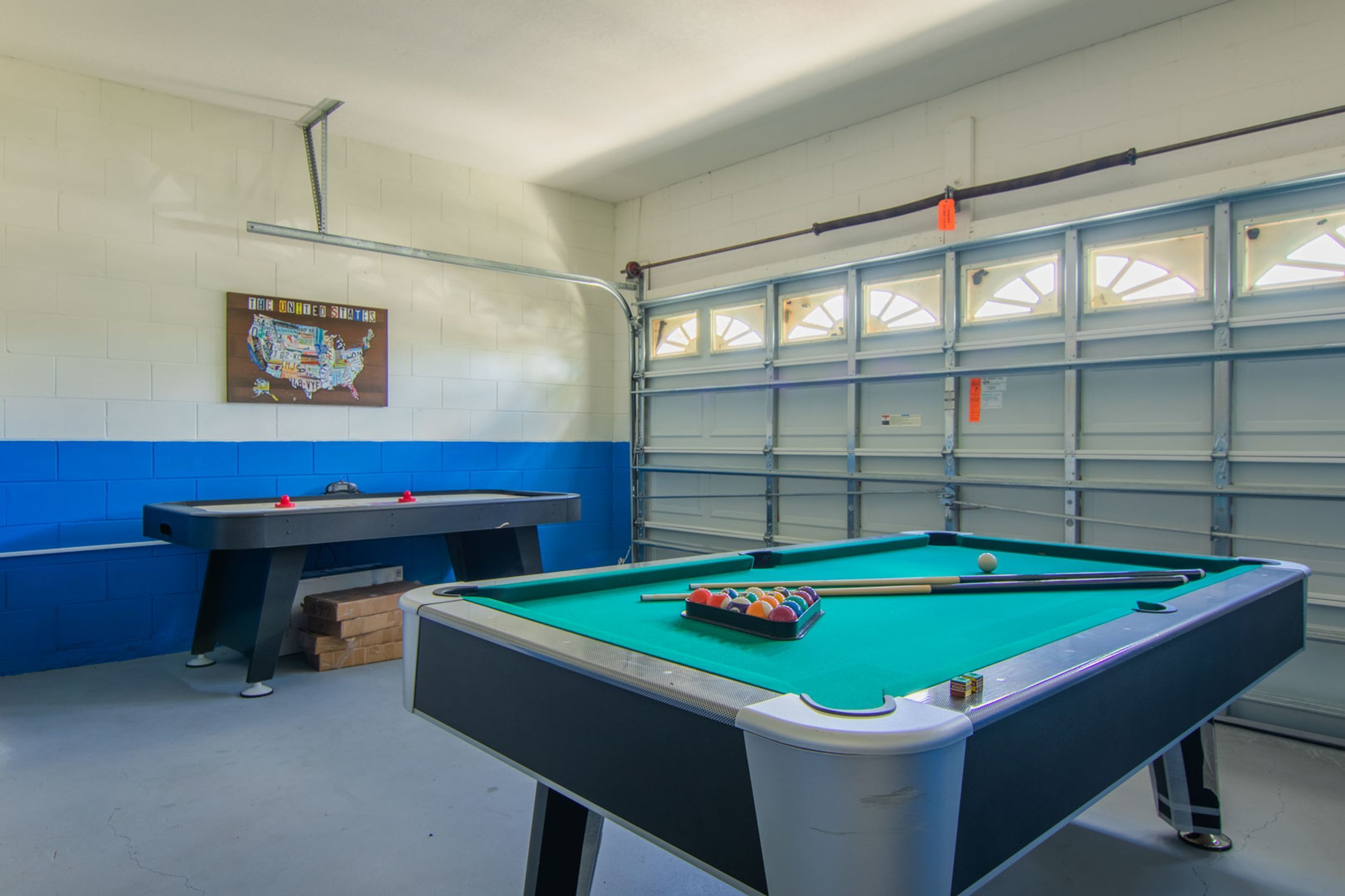 A pool table with a green felt surface, a pool cue, and a set of balls. There is a blue and white wall with a map of the United States.
