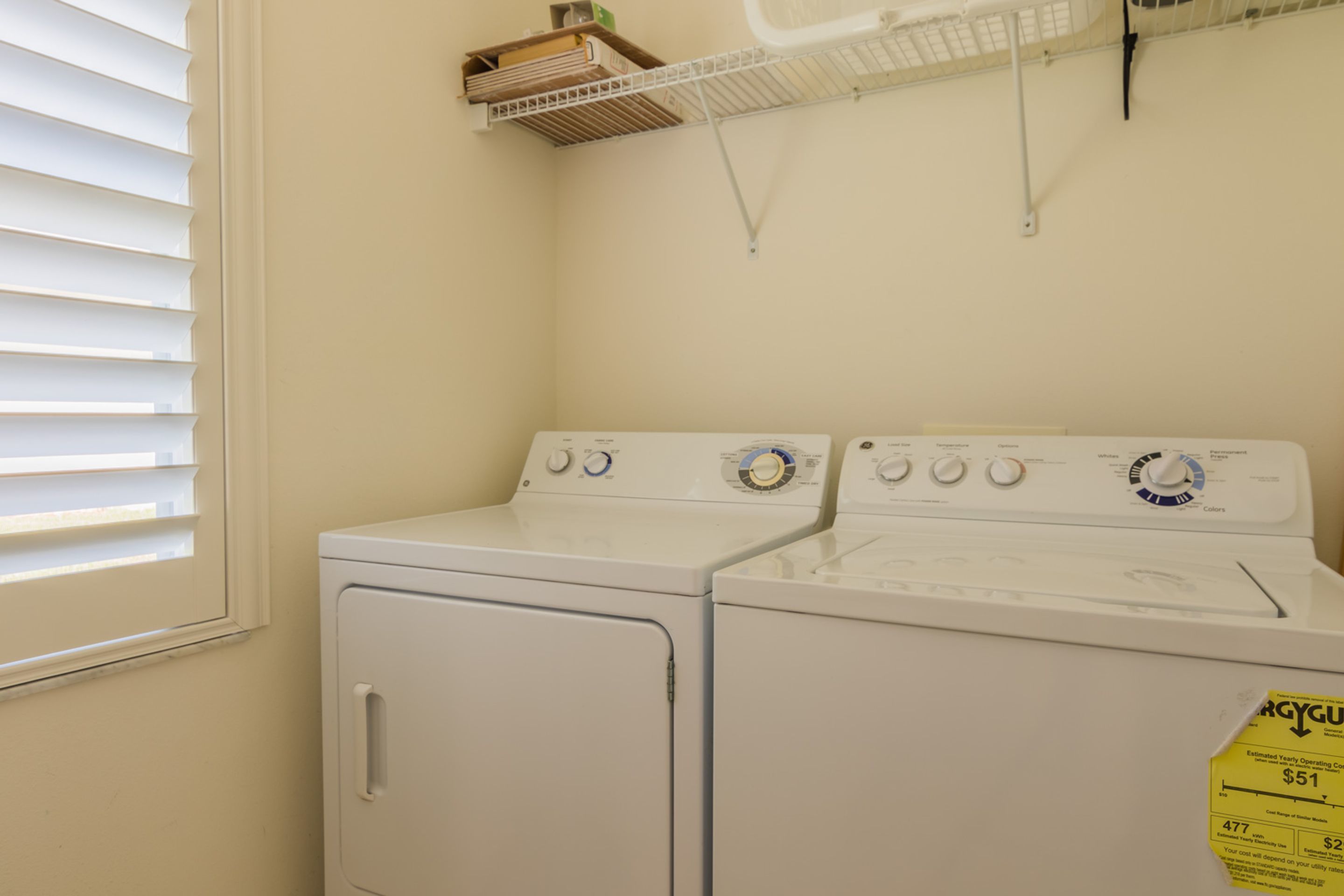 A laundry room with two stacked washers and dryers, a window with blinds, and a shelf above the dryers.