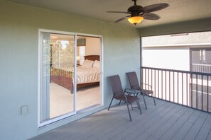 Un balcon avec un ventilateur de plafond, un lit recouvert d’une courtepointe beige et deux chaises.