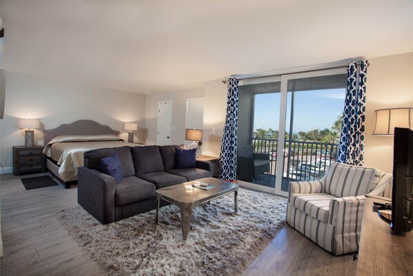 A modern hotel room with a bed, sofa, armchair, and a balcony with a view.