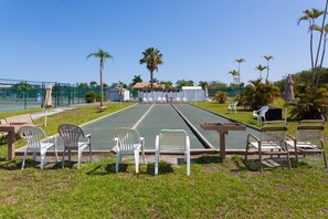 A tennis court with chairs and a table, surrounded by palm trees and a fence.