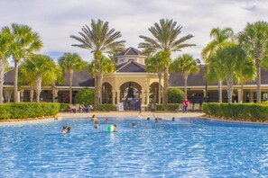 A swimming pool with people swimming and playing, surrounded by palm trees and a building with arches. - Windsor Hills 2591 (Kissimmee)