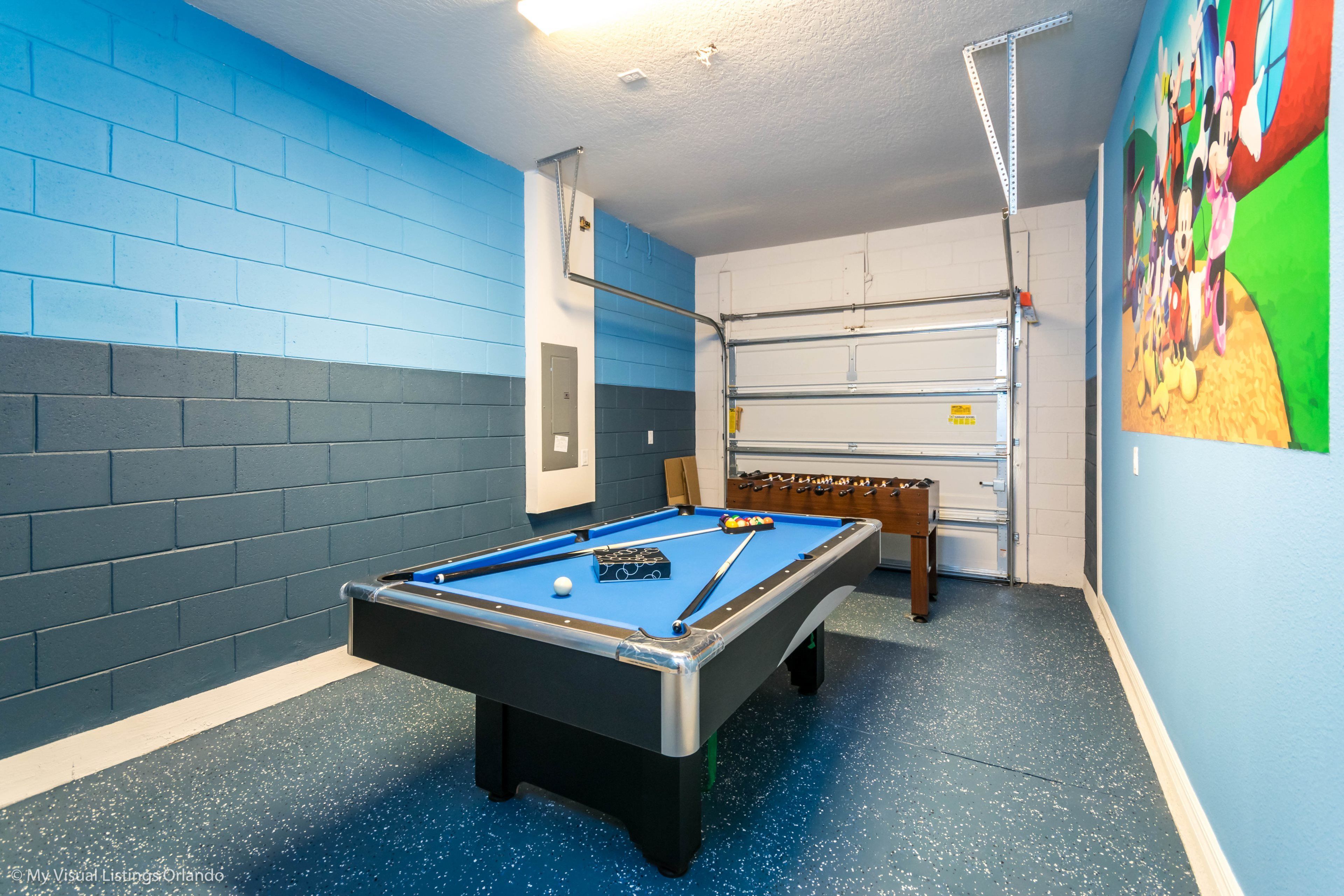 Une table de billard dans une pièce aux murs bleus et gris, avec un ventilateur de plafond et une fresque murale.