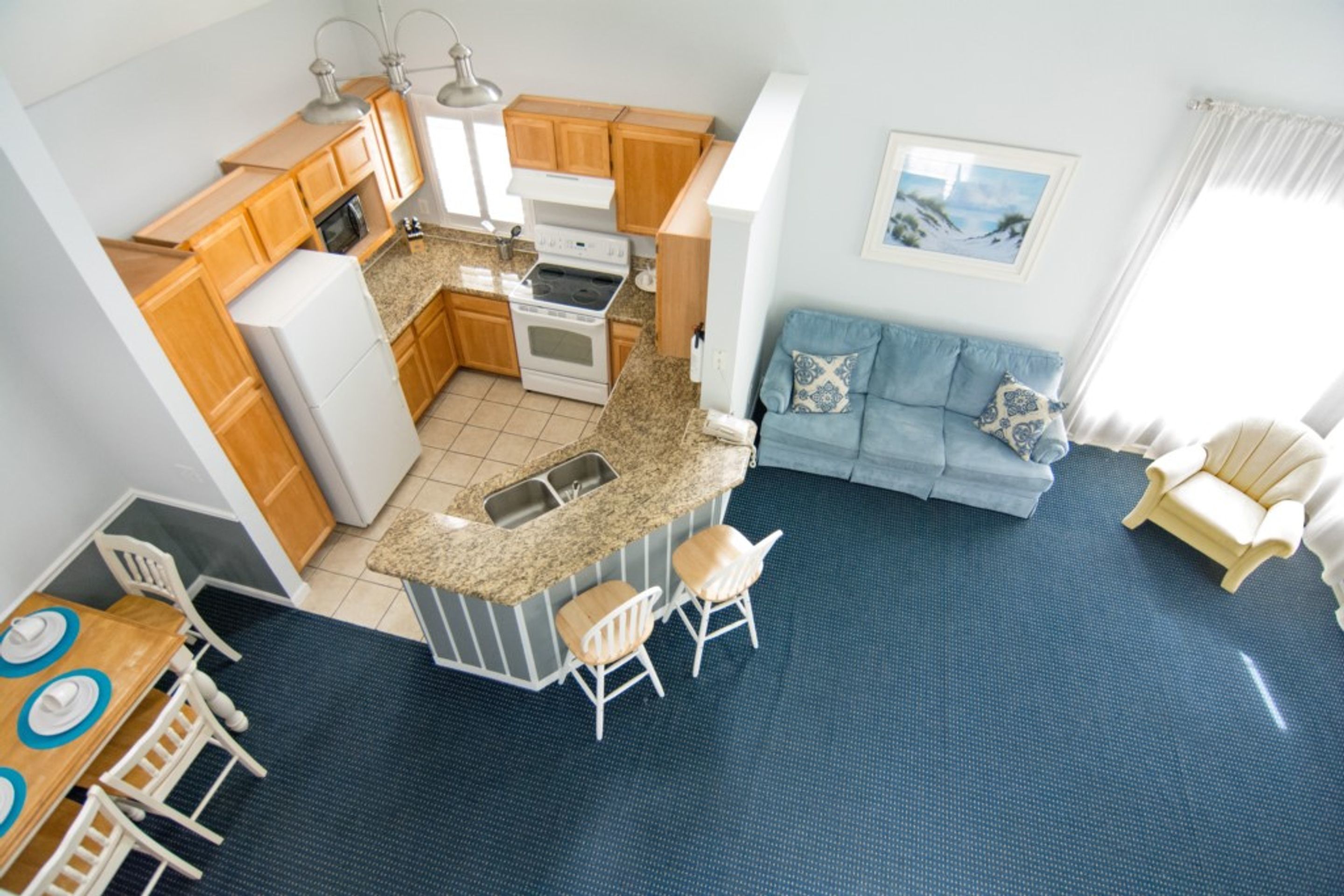 A compact kitchen with wooden cabinets, a white refrigerator, and a granite countertop. There is a blue sofa and a white armchair in the living area.