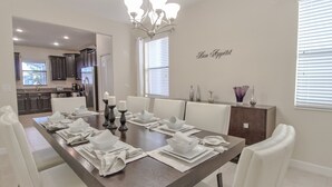 A modern dining room with a long table set for six, white chairs, and a chandelier. - Champions Gate 9033 (Davenport)