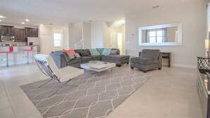 A modern living room with a sectional sofa, a white coffee table, and a grey rug. There is a kitchen area with dark cabinets and a microwave. - Champions Gate 9033 (Davenport)