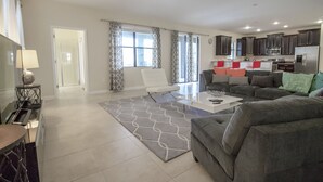 A modern living room with a grey sofa, a coffee table, and a patterned rug. There is a kitchen area with dark cabinets and a refrigerator. - Champions Gate 9033 (Davenport)