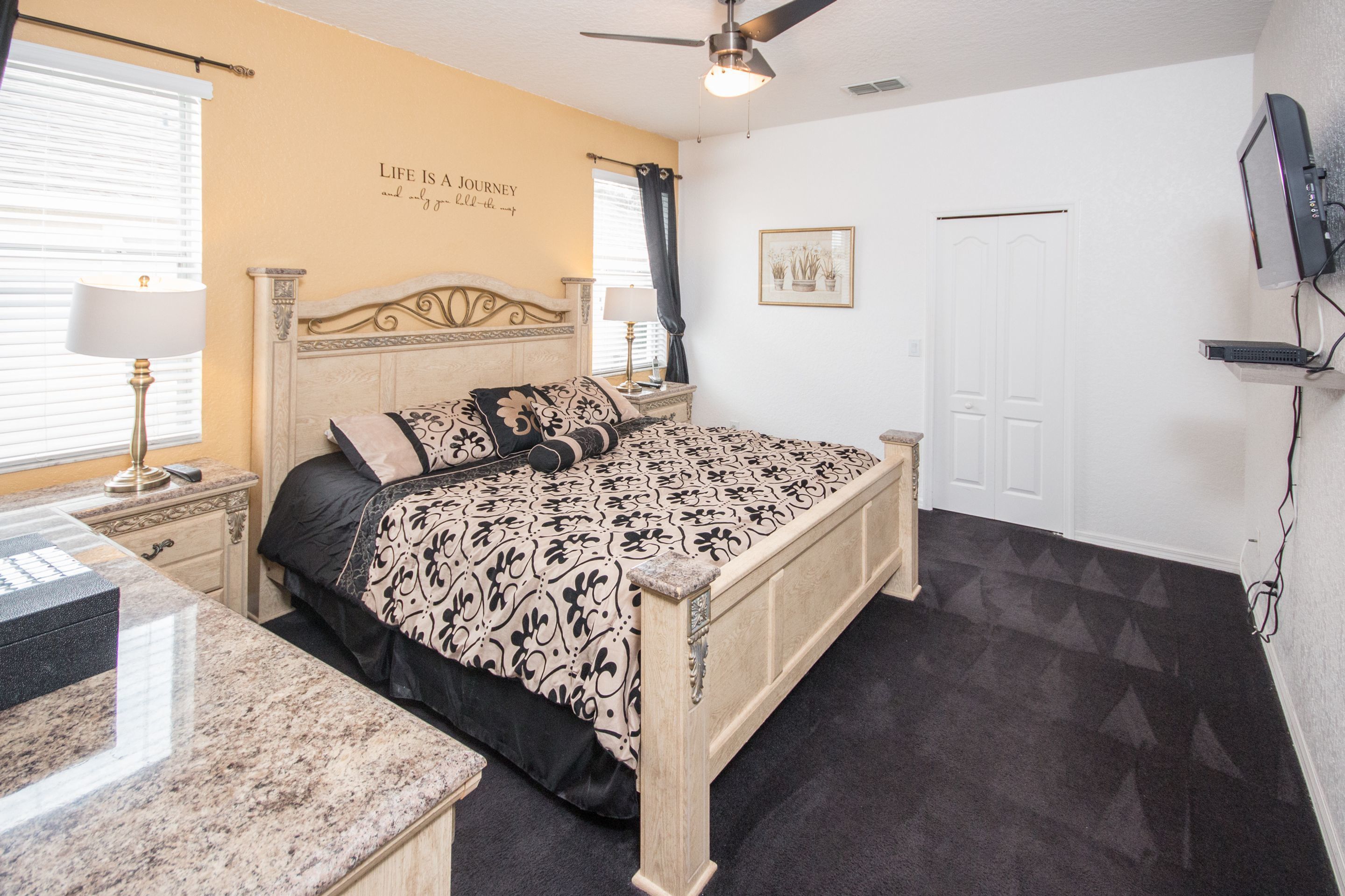 A bedroom with a bed, nightstands, a desk, a ceiling fan, a TV, and a door.