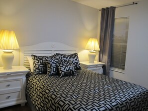 A neatly arranged bedroom with a patterned bedspread, two bedside lamps, and a window with blinds.