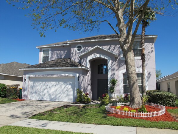 A two-story house with a garage, a front yard with a tree and a white picket fence, and a clear blue sky.