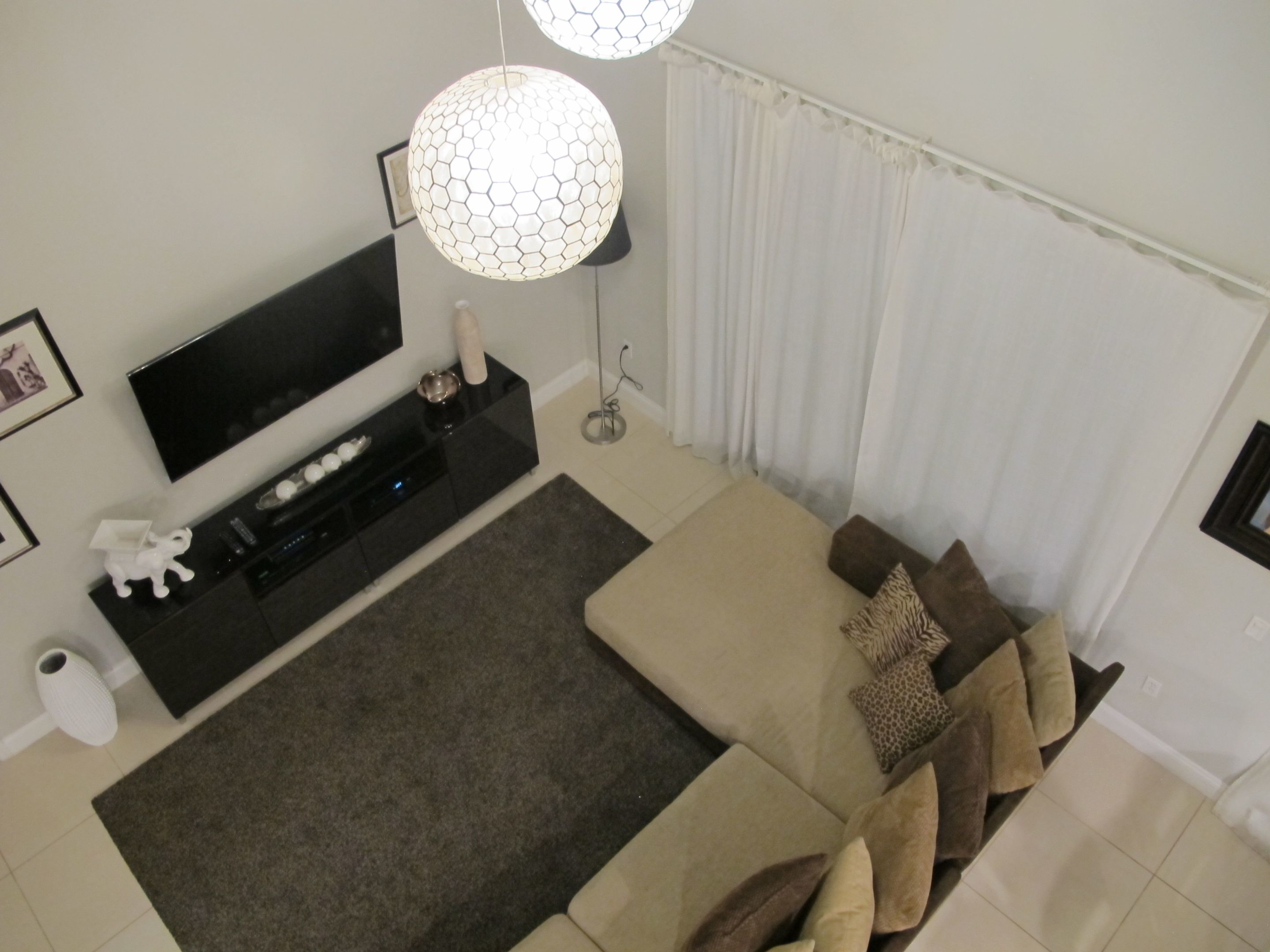 A modern living room with a flat-screen TV, a black entertainment unit, a beige sofa with multiple cushions, a grey rug, and a white curtain.