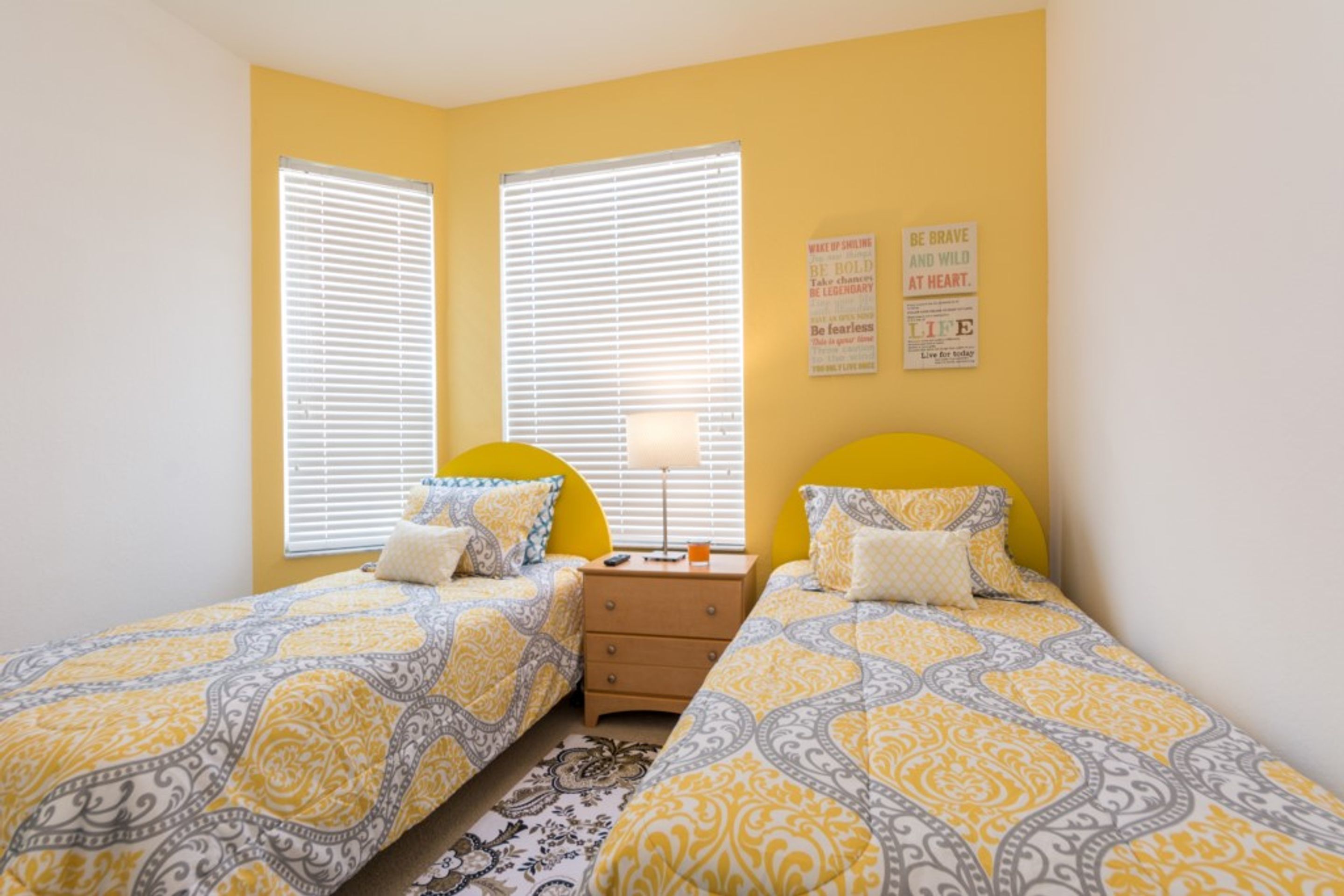 Quarto com duas camas, cada uma com roupa de cama estampada, mesa de cabeceira com abajur e janela com persianas.