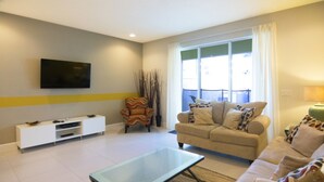 A living room with a sofa, a glass coffee table, a TV mounted on the wall, and a window with curtains. - Serenity 17417 B (Clermont)