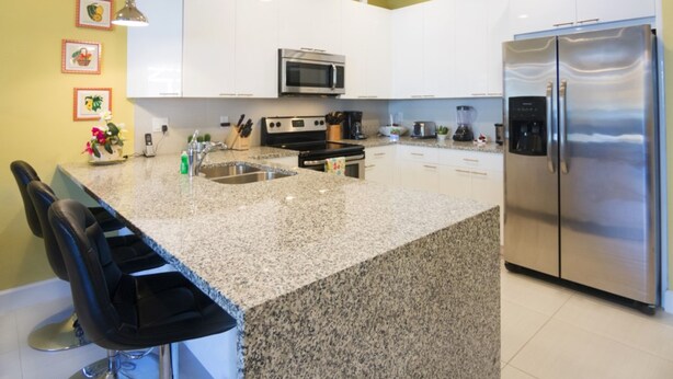 A modern kitchen with white cabinets, a stainless steel refrigerator, granite countertops, and a built-in oven. - Serenity 17417 B (Clermont)
