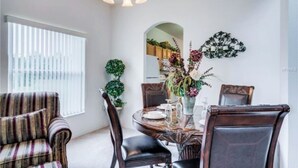 Un coin repas avec une table ronde, des chaises et un lustre. Il y a une fenêtre avec des stores et une plante sur le côté.