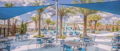 An outdoor patio area with tables and chairs, palm trees, and a pool in the background.