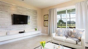 A modern living room with a flat-screen TV mounted on a wooden wall, a white sofa with patterned cushions, and a coffee table with a vase of yellow flowers.