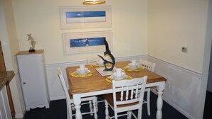 A dining area with a wooden table and white chairs, framed artwork on the wall, and a small cabinet to the left.