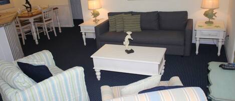 Uma sala de estar com um sofá cinzento, poltronas listradas, uma mesa de centro branca e duas mesas de cabeceira com abajures.