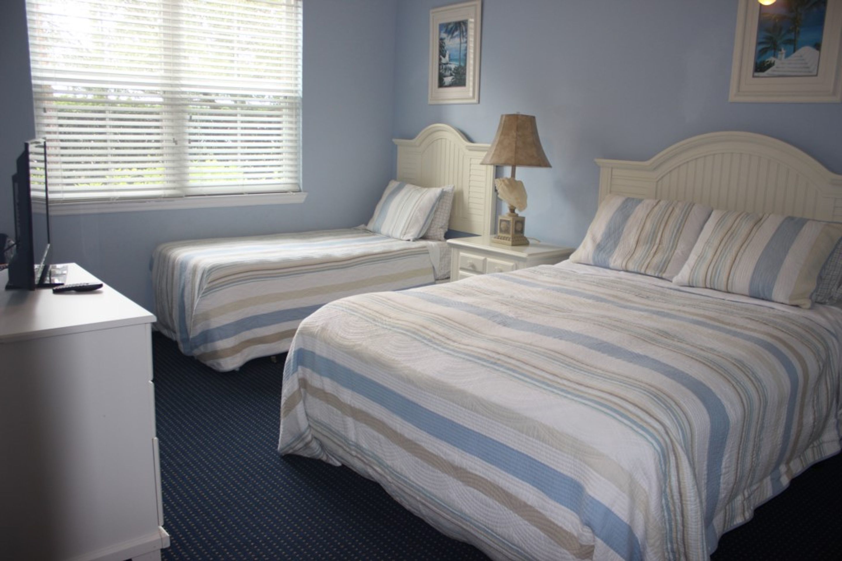 A hotel room with two single beds, a nightstand with a lamp, a dresser, and a window with blinds.