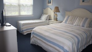 A hotel room with two single beds, a nightstand with a lamp, a dresser, and a window with blinds.