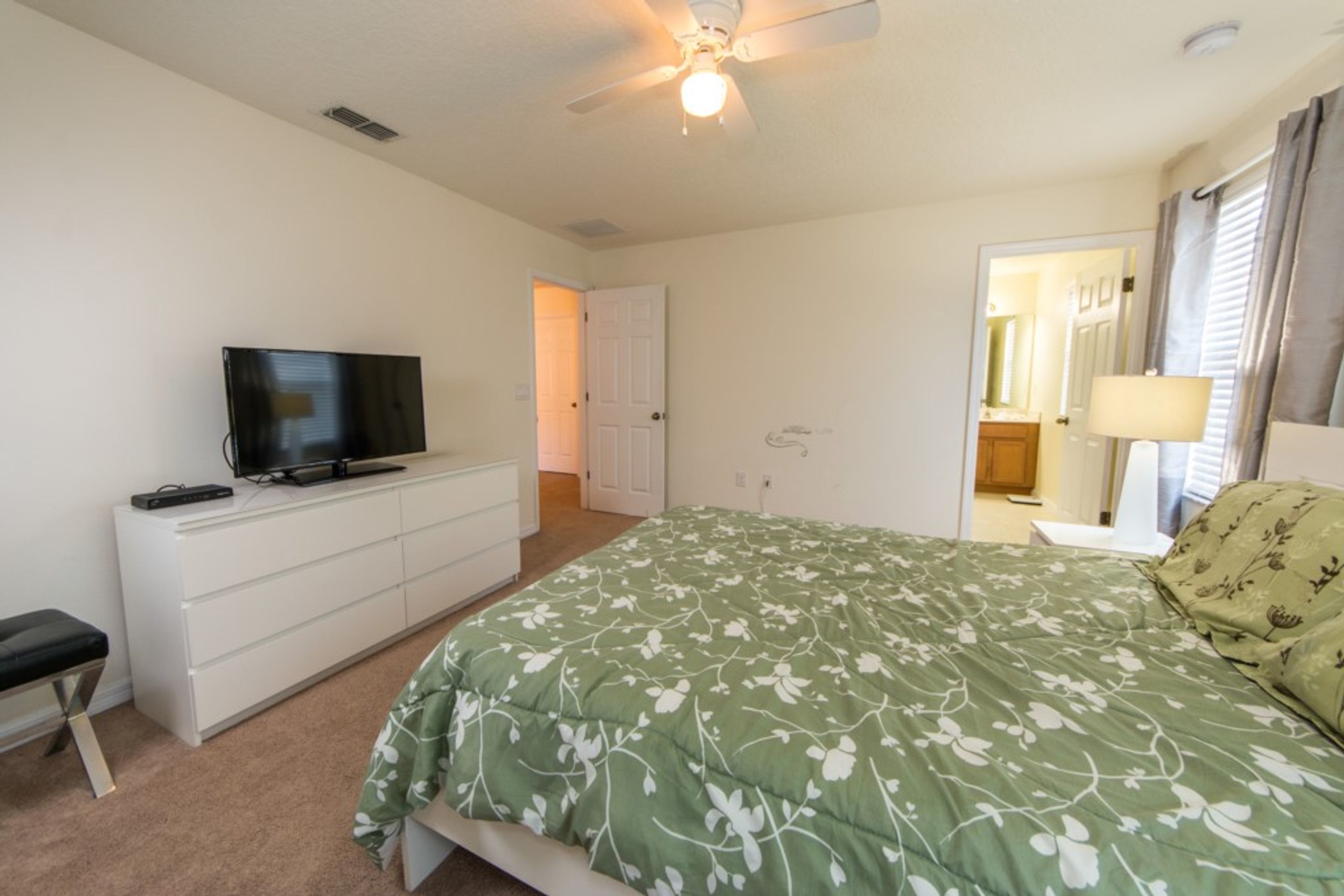 Une chambre à coucher avec un lit, un téléviseur posé sur une commode, un ventilateur de plafond et une porte donnant sur une autre pièce.