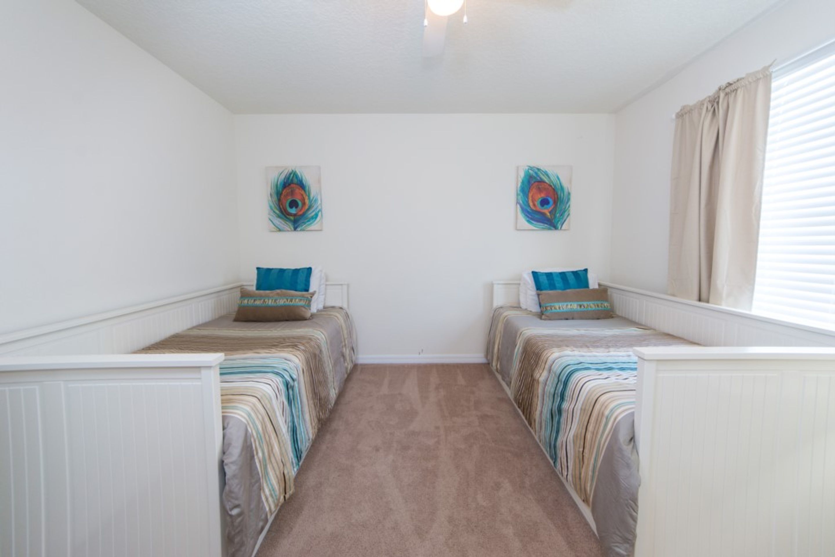 A bedroom with two single beds, a carpeted floor, and two wall-mounted peacock-themed paintings.