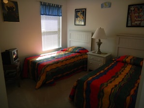A bedroom with two beds, a nightstand, a lamp, a window with blinds, and a television.