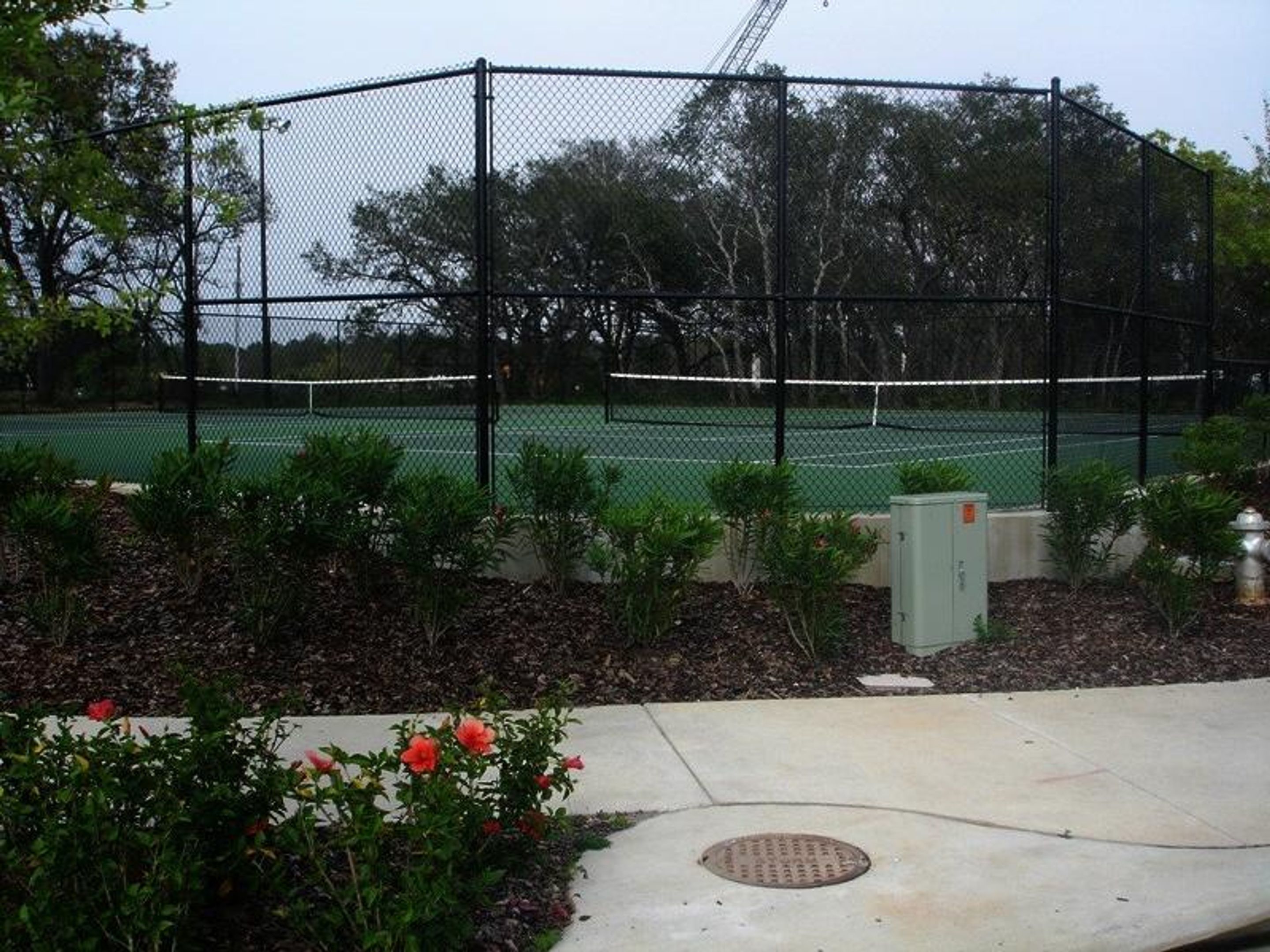 A tennis court surrounded by a chain-link fence, with a manhole cover and some greenery in the foreground.