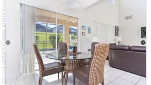 A dining area with a glass table and wicker chairs, a brown leather sofa, and a view of a green landscape through large glass doors. - Emerald Island Resort 8622 (Kissimmee)