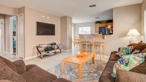 A living room with a sofa, a coffee table, and a rug. There is a kitchen area in the background with cabinets and a refrigerator. - Southern Dunes 1120 (Haines City)